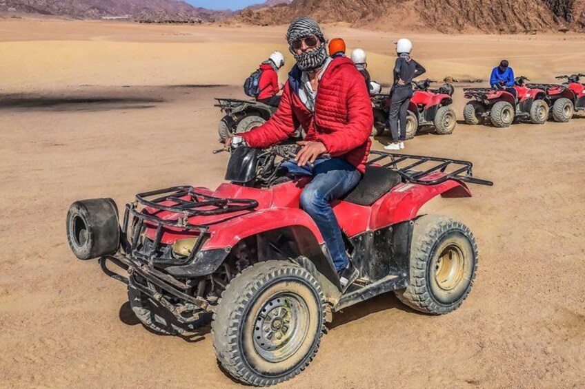 A person riding a red quad bike in a desert landscape with sand dunes and other quad bikes in the background. 4