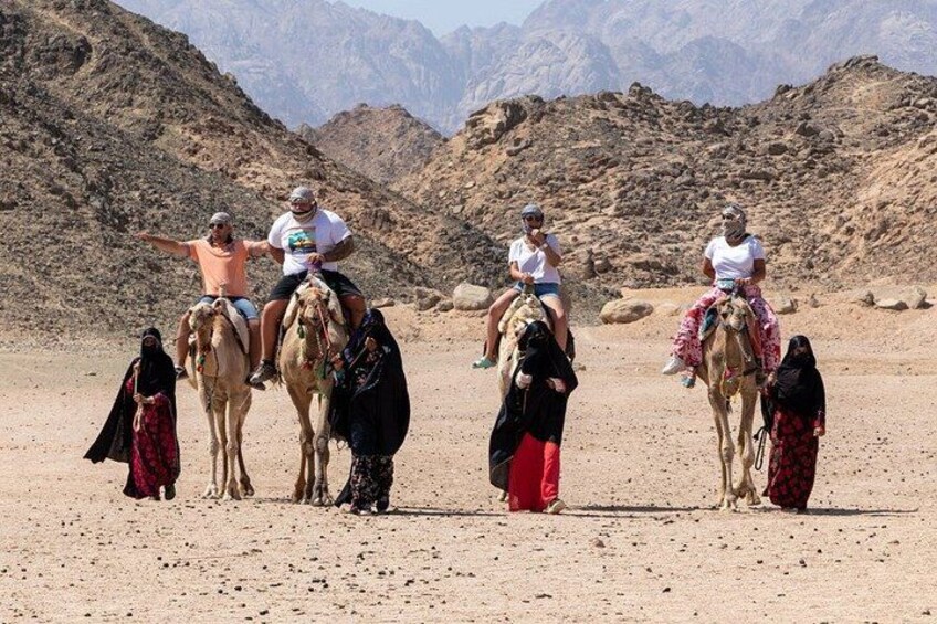 A group of people riding camels in a desert landscape with rocky hills in the background. 4