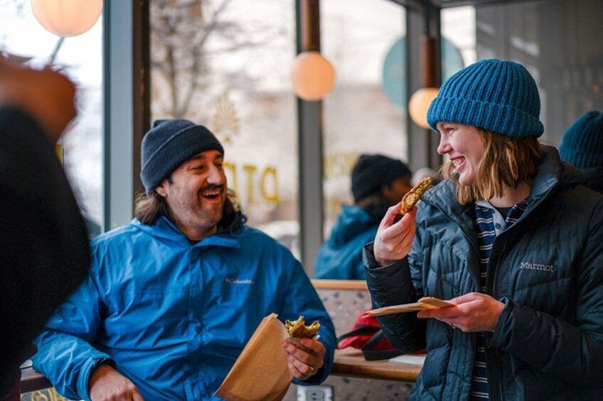 Two people eating at an outdoor cafe, one holding a sandwich and the other a drink. 3