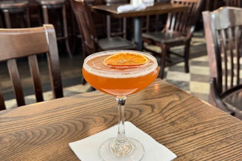 A cocktail in a stemmed glass with an orange garnish on a wooden table in a restaurant. 5