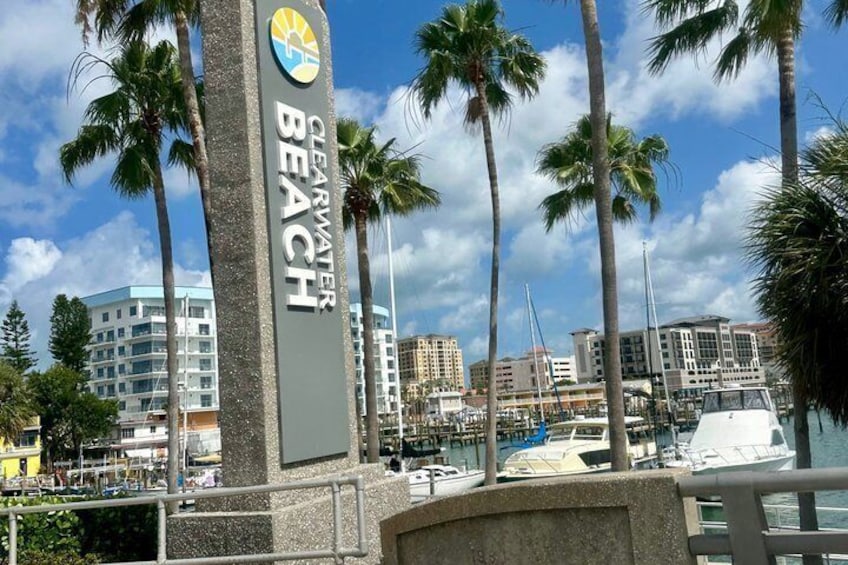 A coastal cityscape with a prominent sign for Clearwater Beach, palm trees, and a marina with boats. 4