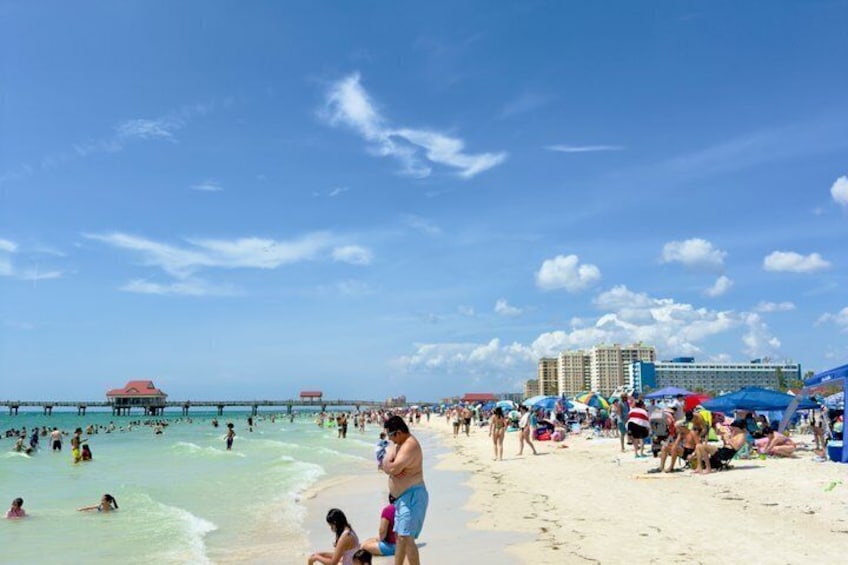 A crowded beach with people swimming and sunbathing, a pier extending into the water, and a hotel in the background. 1