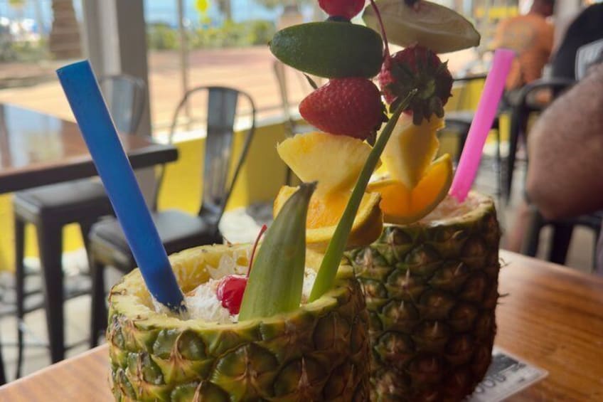 Two drinks in pineapple cups with fruit garnishes and straws. 2