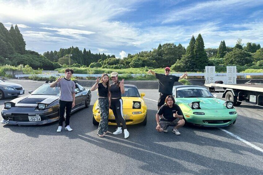 A group of people posing with sports cars in a parking lot. 1