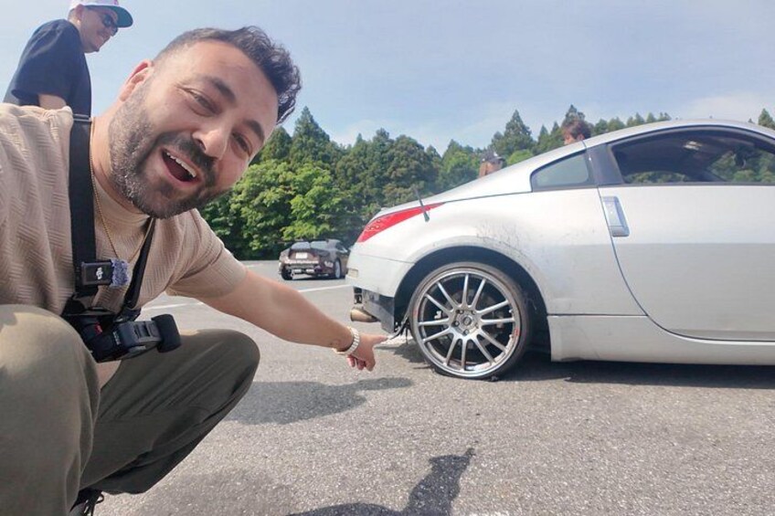 A person crouching next to a white car with a camera on their shoulder. 4
