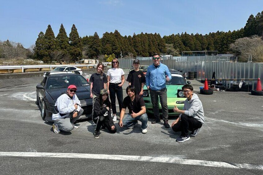 A group of people posing with two cars in a parking lot. 5