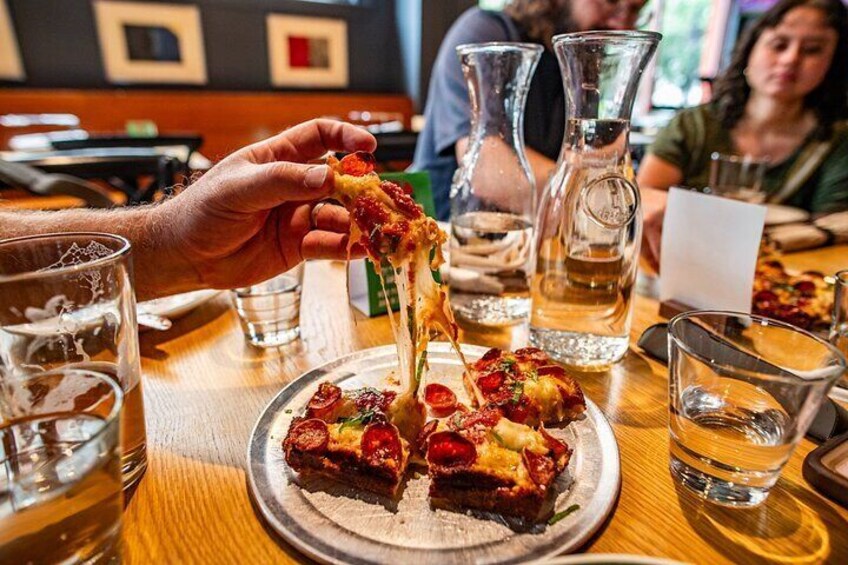 A person holding a slice of pizza with toppings, a plate with two slices, and glasses of water on a wooden table. 1