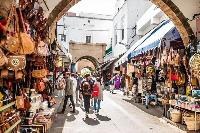A bustling market street with people walking, various stalls selling bags and textiles, and an archway in the background. 4