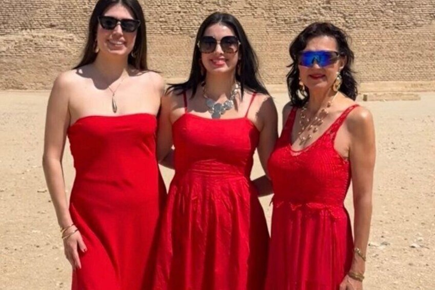 Three women in red dresses standing in a desert with a large stone structure in the background. 4