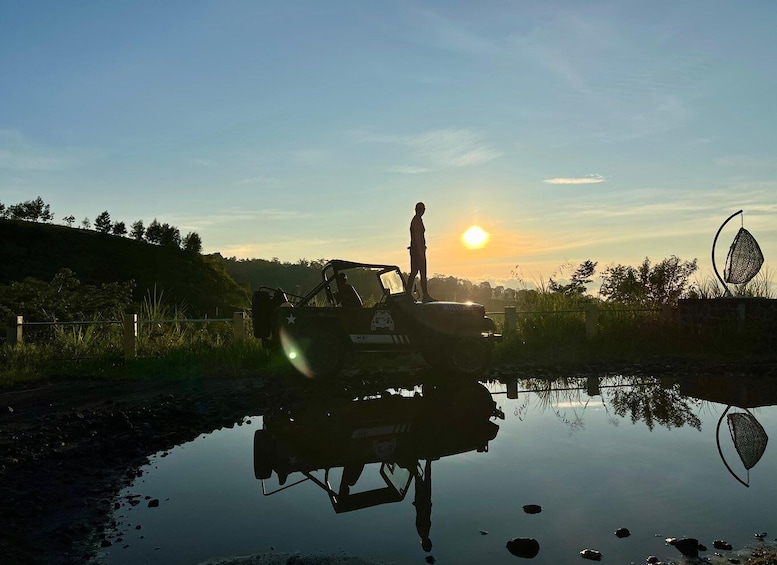 Ett fordon på en vattenyta som speglar omgivningen, med en person stående på det. I bakgrunden syns en solnedgång, och på vänster sida finns ett staket med växtlighet. 5