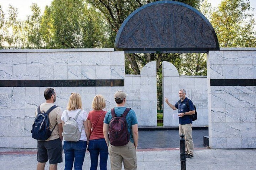 A group of people standing in front of a memorial wall with inscriptions and a large archway. 3