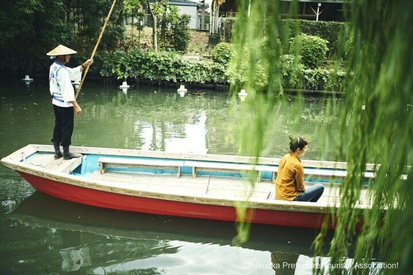 A person in a boat on a calm river, another person seated inside, and a person standing on the shore with a pole. 1