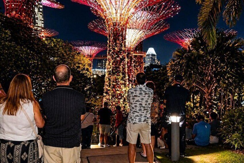 A nighttime outdoor scene with illuminated structures resembling trees, a city skyline in the background, and people walking around. 1