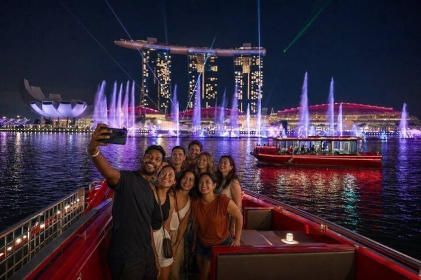 A group of people on a boat with a city skyline and illuminated fountains in the background. 1
