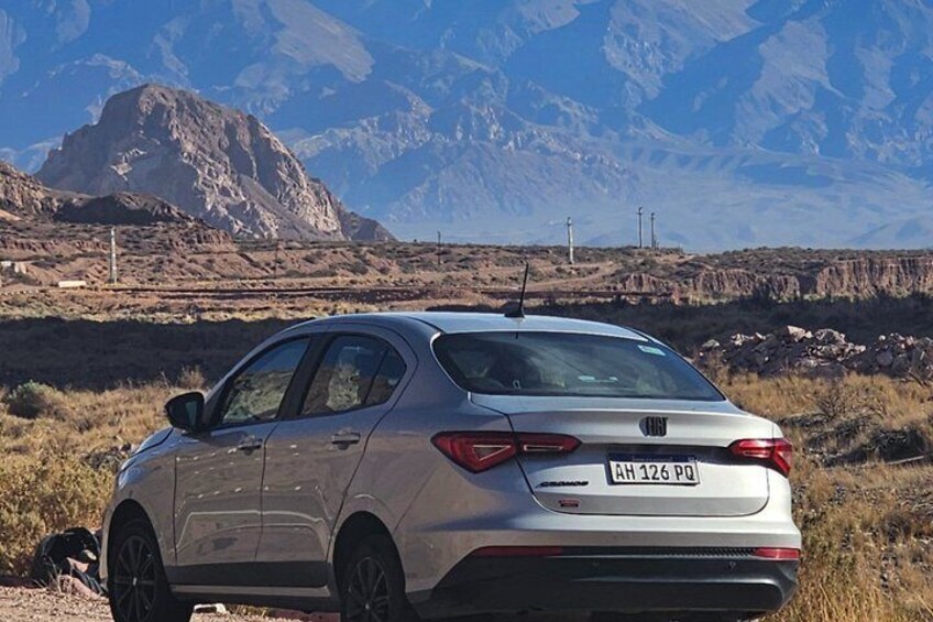 A silver car parked on a dirt road with mountains in the background. 5