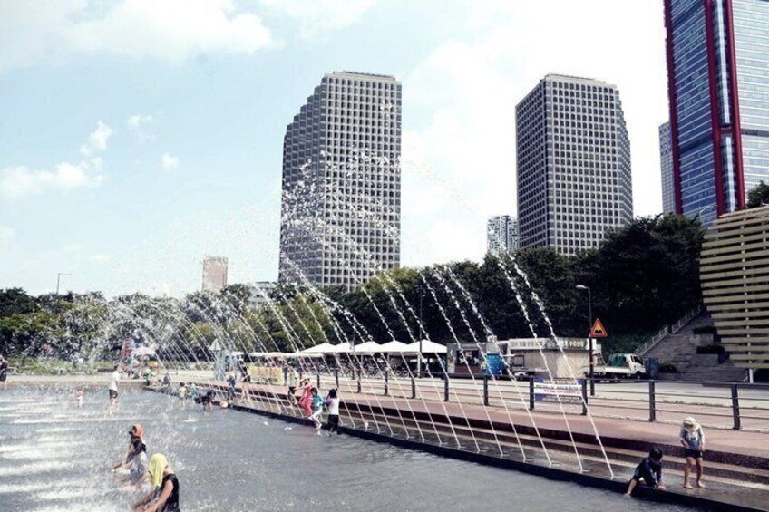 A water fountain feature with water jets, a pedestrian walkway, and modern buildings in the background. 2