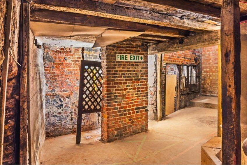 A brick corridor with a fire exit sign, wooden beams, and a metal gate. 4