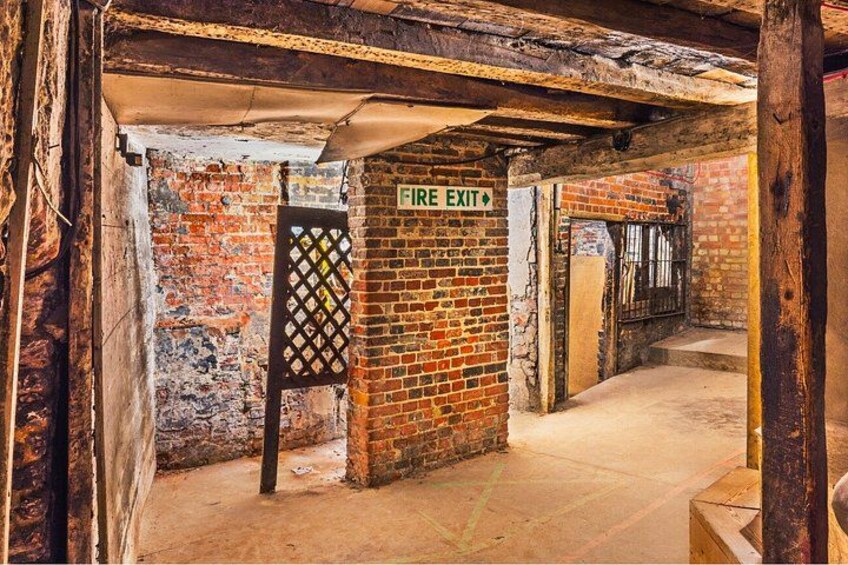 A dimly lit brick corridor with a fire exit sign, a metal gate, and a staircase leading to a higher level. 2