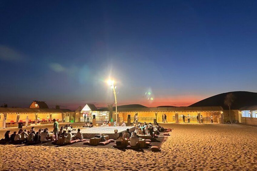 A beach with people sitting and standing, a building with a thatched roof, and a sunset in the background. 4