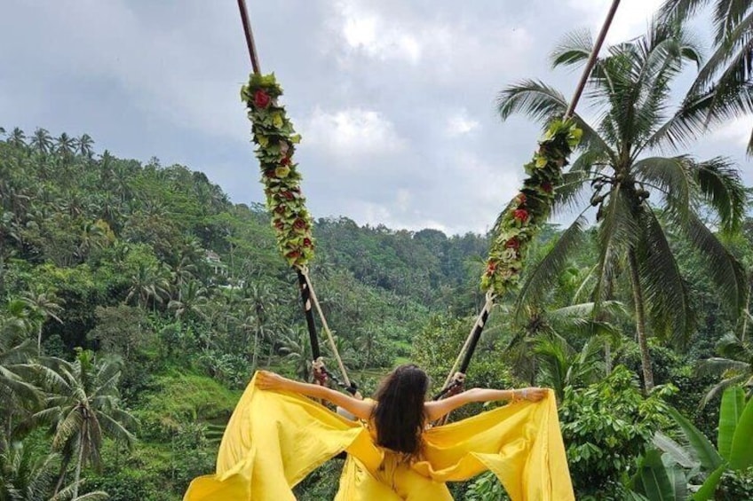 A person in a yellow dress swinging on a rope with floral decorations, surrounded by a lush tropical landscape. 4