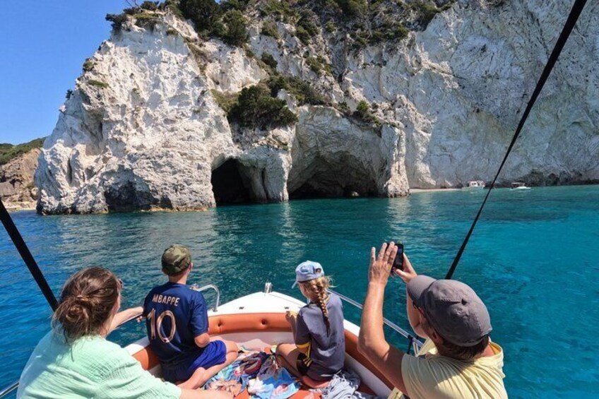 A boat with people taking photos, a rocky coastline, and a clear blue sea. 1