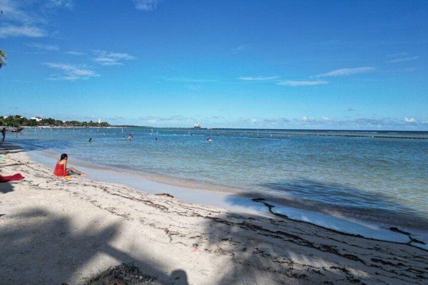 A beach with clear blue water, a person sitting on the sand, and a distant shoreline with buildings. 3