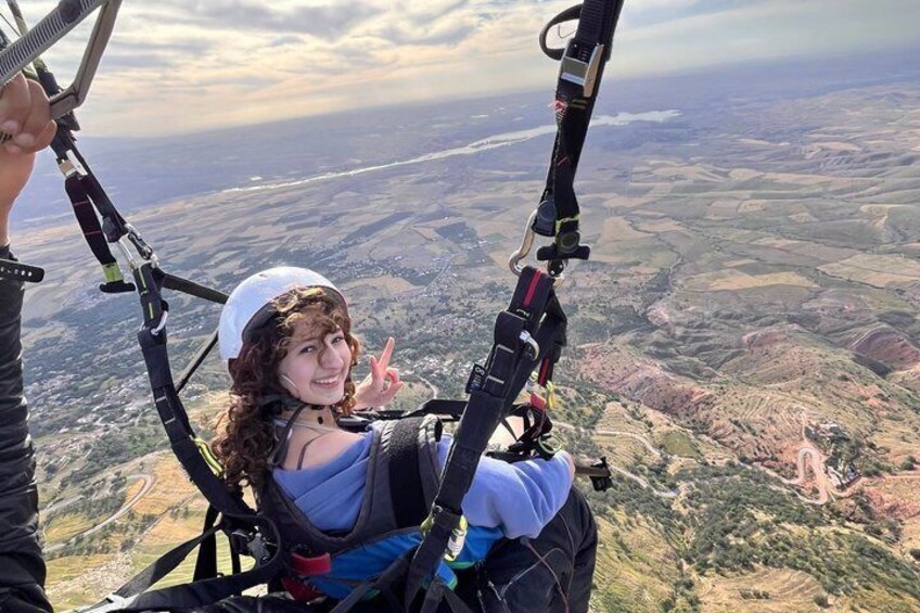 A person in a harness with a parachute, holding onto the harness lines, with a landscape of fields and hills in the background. 3
