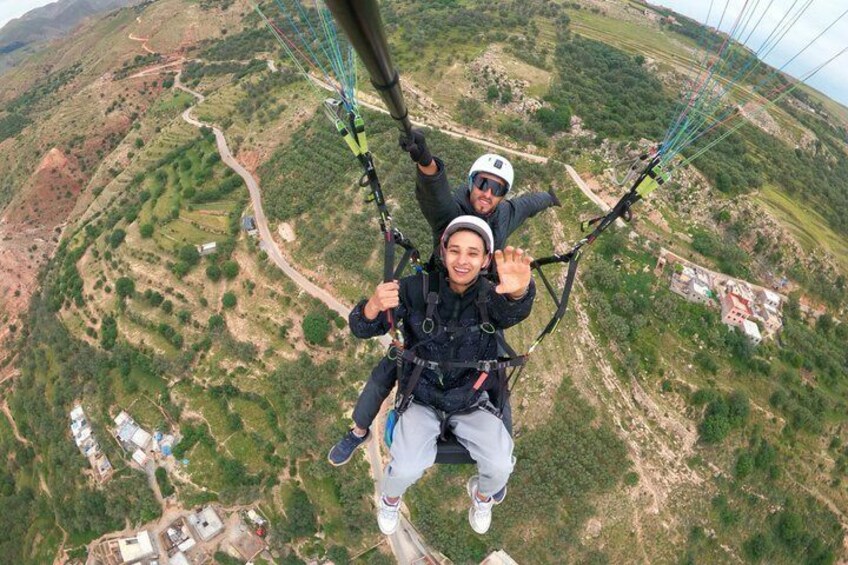 A person paragliding with a guide, over a mountainous landscape with terraced fields. 1