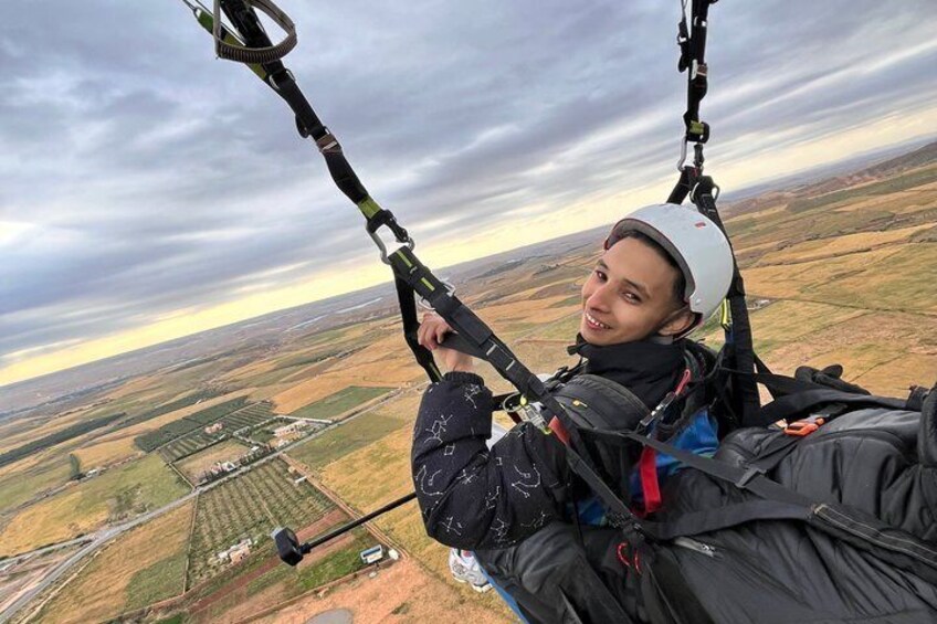 A person paragliding with a harness and helmet, over a rural landscape with fields and a road. 5