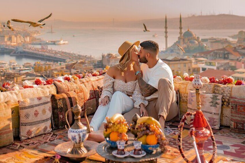 A couple sitting on a decorated terrace overlooking a cityscape with a mosque and waterfront. 2