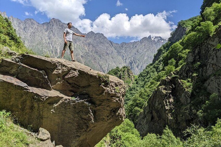 A person standing on a rock ledge with mountains in the background. 1