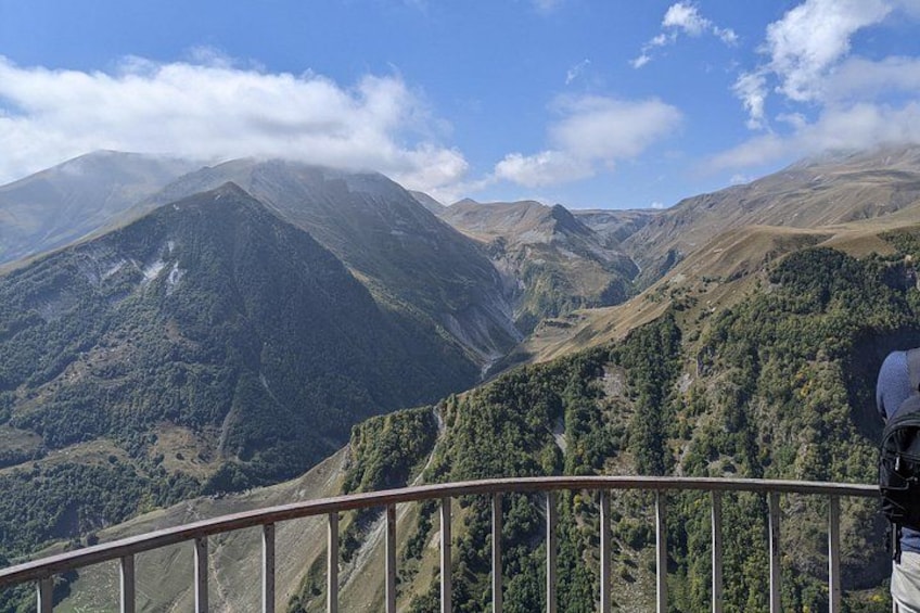 A mountainous landscape with a clear sky, a person standing on a railing, and a forested valley. 5