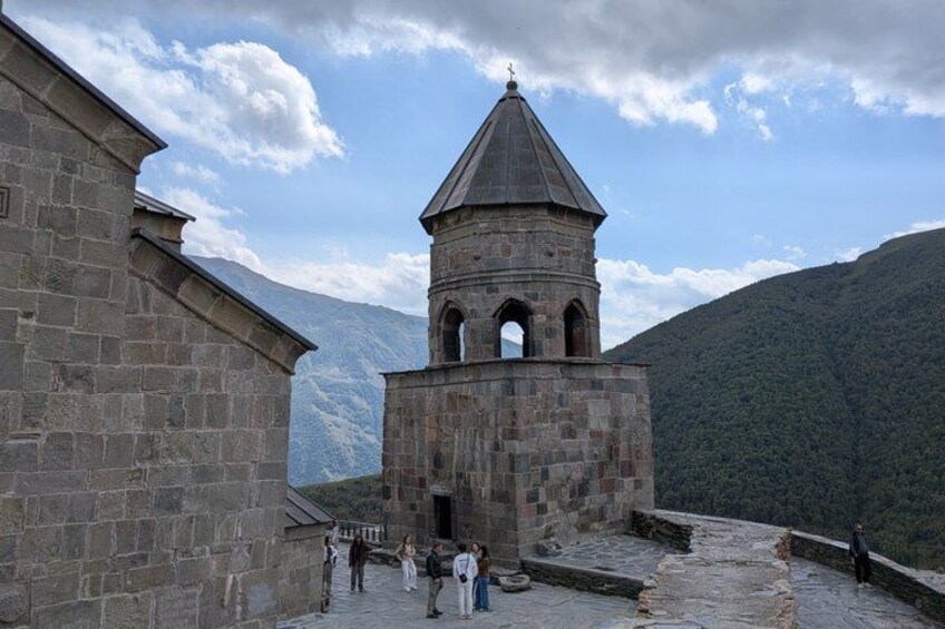 A stone tower with a conical roof, a stone building, and a mountainous landscape in the background. 2