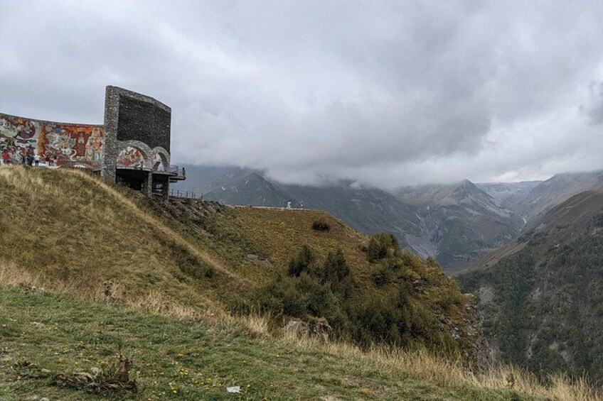 Een bergachtig landschap met een gebouw versierd met kleurrijke muurschilderingen, een grasheuvel en een vallei. 3