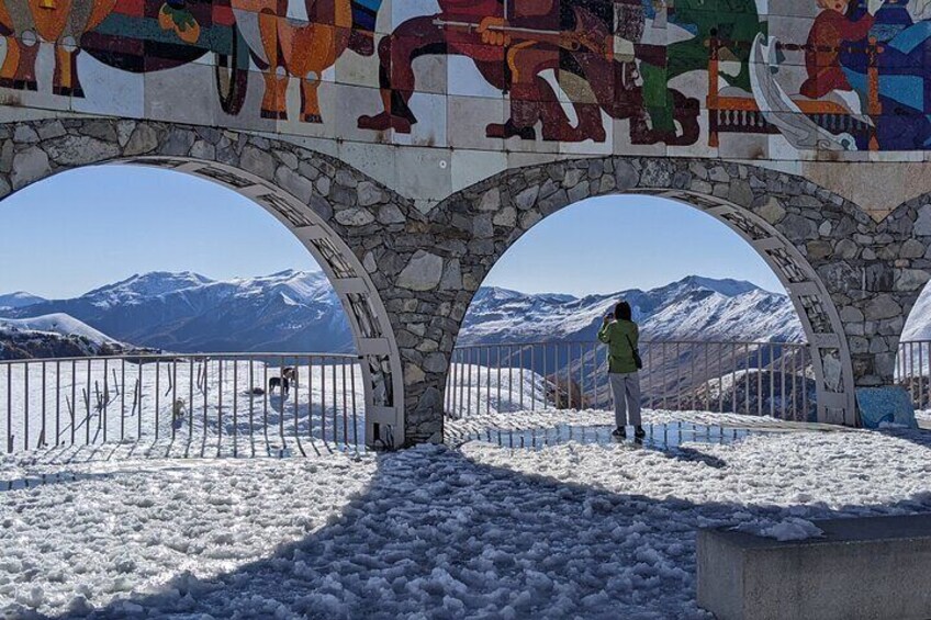 A person standing under a colorful mural archway with snow-covered mountains in the background. 3