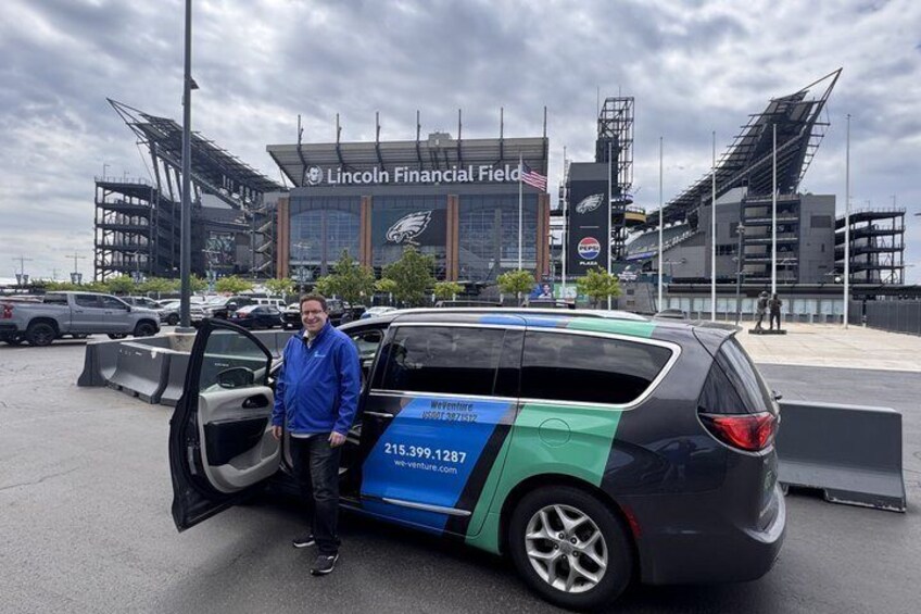 A man standing next to a car with a green and blue advertisement on the side, parked in front of Lincoln Financial Field. 1