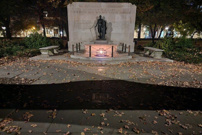 A monument with a statue, surrounded by fallen leaves and benches, illuminated by artificial lighting. 1