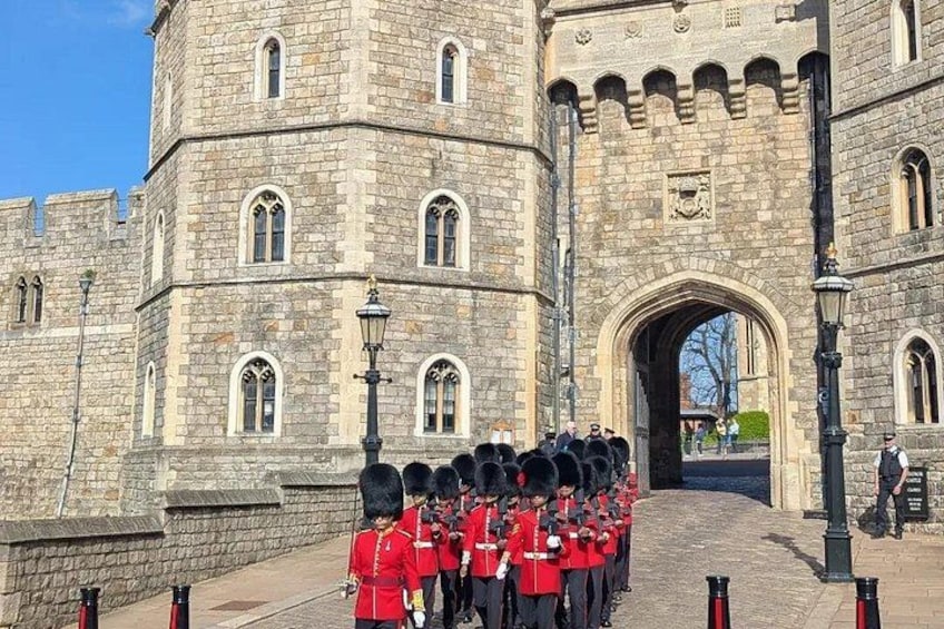 A historic stone building with a large arched entrance, a group of guards in red uniforms, and a clear blue sky. 3