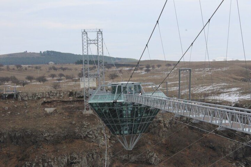 A suspension bridge with a green structure, metal framework, and a metal tower in a rural landscape. 1