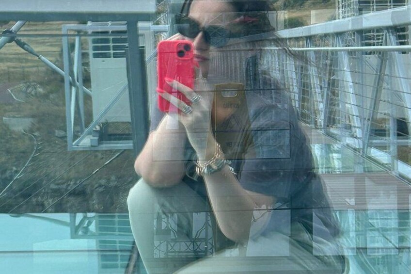 A person taking a selfie with a red phone, wearing a watch, and sitting in front of a glass window with a metal structure in the background. 3