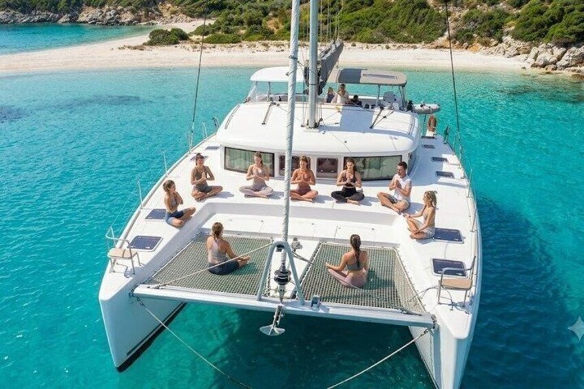 A white catamaran with people meditating on deck, surrounded by turquoise water and a sandy beach. 1