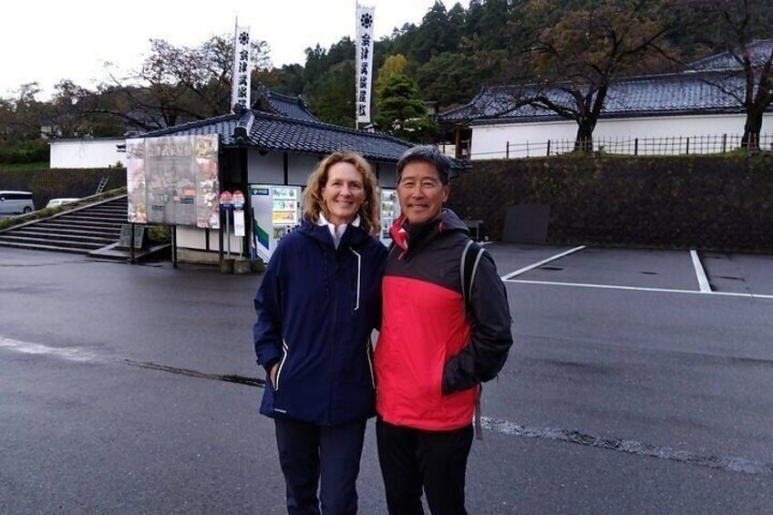 Two people standing in front of a traditional Japanese building with a tiled roof and a small shrine. 3