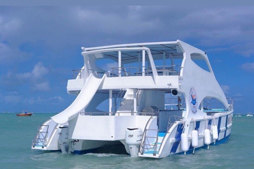 A large white boat with a covered upper deck and a single white sail, floating on calm blue water. 1