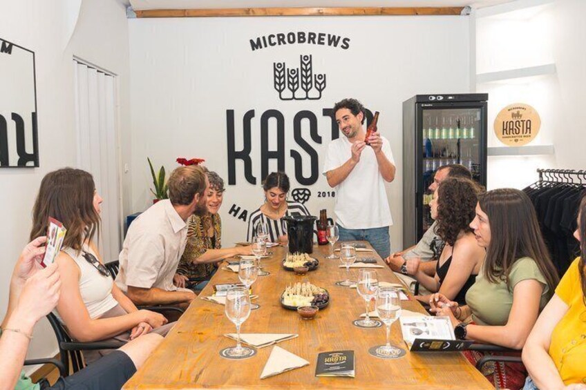 A group of people seated around a wooden table with a bottle of wine, glasses, and a cake in a room with a wall sign reading 'MICROBREWS KAST 2018'. 1