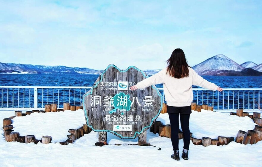 A person standing in front of a wooden sign with text, overlooking a snowy landscape and a body of water with mountains in the distance. 1