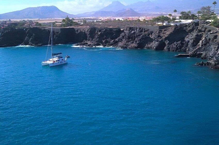 A sailboat on a calm blue sea with a rocky coastline and mountains in the background. 2