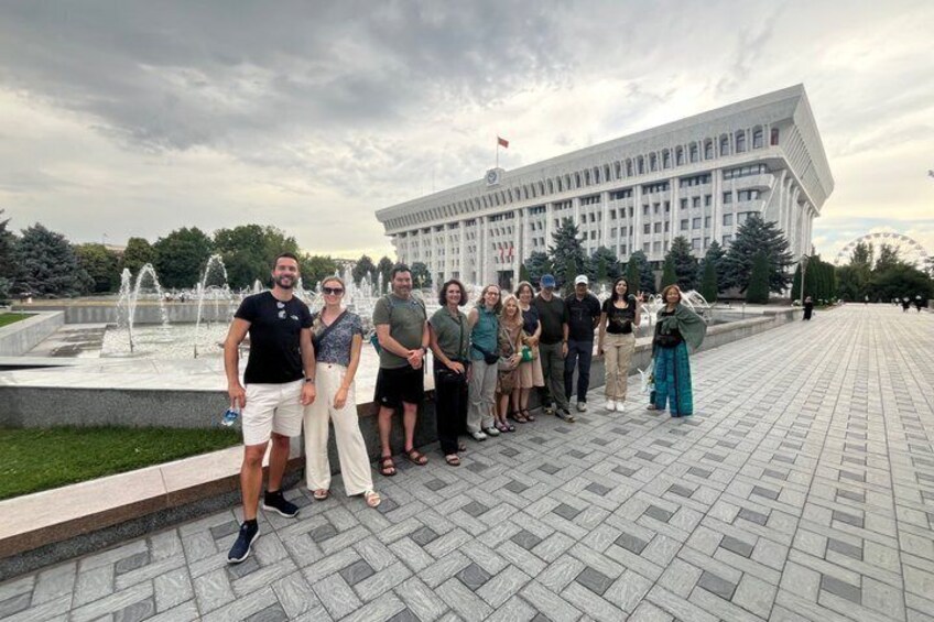 A group of people standing in front of a fountain and a large building with a flag on top. 1