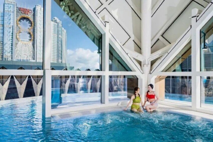 An indoor swimming pool with a glass ceiling, a cityscape view, and two people sitting by the pool. 1