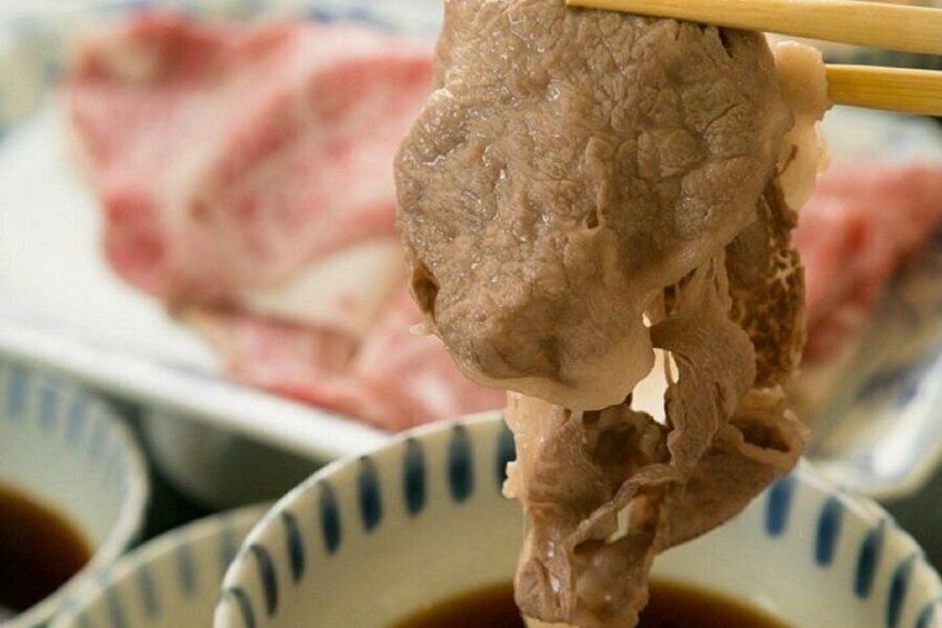 A piece of meat being held by chopsticks over a bowl, with a plate of sliced meat in the background. 2