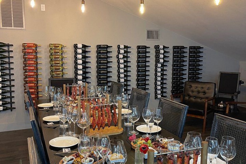 A wine tasting room with rows of wine bottles, a long table set for tasting, and a cozy seating area. 2